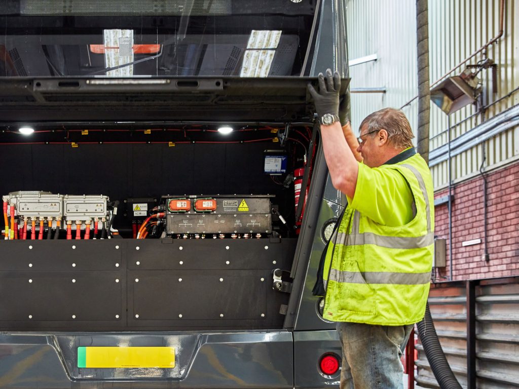 A technician in a high-visibility vest and gloves is inspecting the battery compartment of an electric bus. The bus's rear hatch is open, revealing multiple battery modules and electrical components with various colored wires and connectors. The setting is a bus depot with electrical equipment visible in the background.