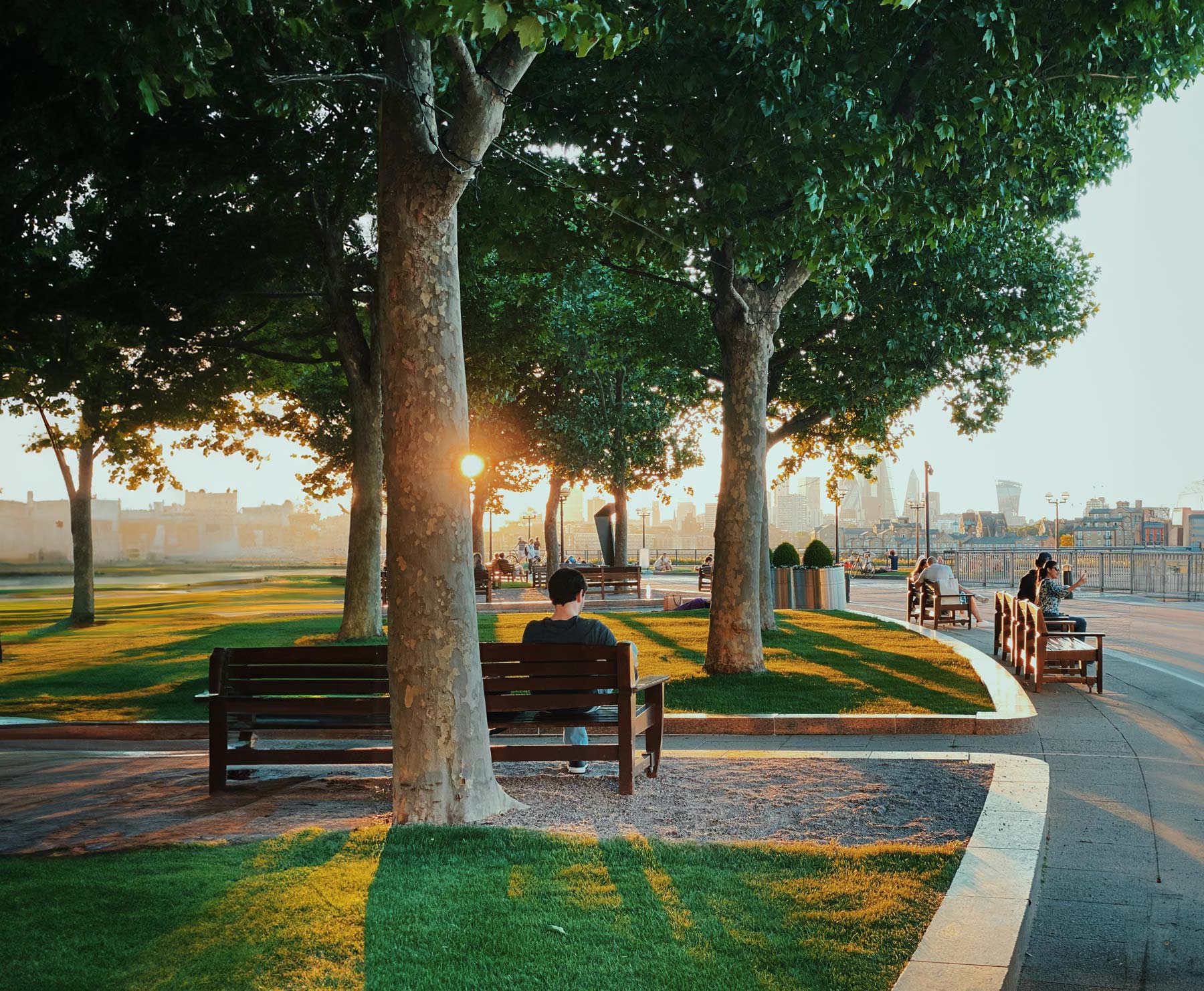 People sitting on benches in a park with trees and well-maintained grass, enjoying a sunset with the sun casting long shadows. The background shows a city skyline with various buildings and structures.