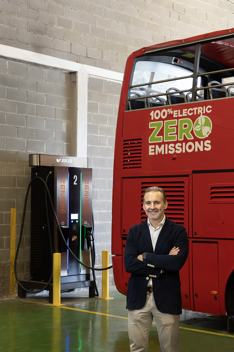 Grupo Julià CEO standing in front of a red electric double-decker bus with “100% Electric Zero Emissions” branding, next to a Zenobē charging station inside the Hospitalet de Llobregat depot in Barcelona. The image highlights the partnership between Grupo Julià and Zenobē to electrify the city tour fleet.