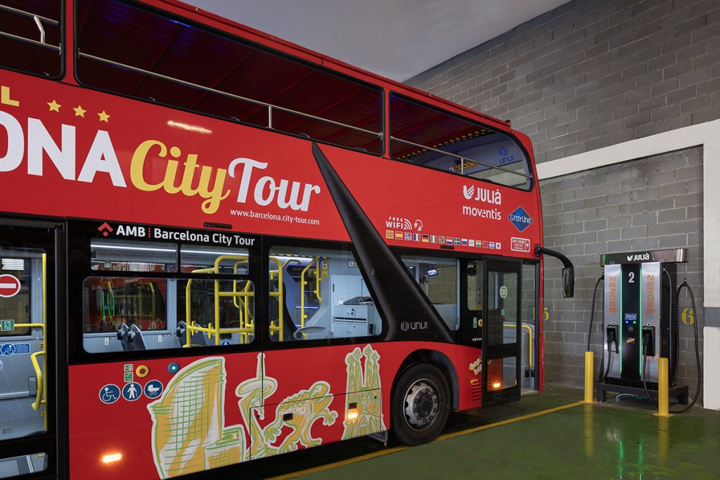 A red double-decker Barcelona City Tour electric bus parked next to a Zenobē-branded charging station inside a bus depot in Hospitalet de Llobregat, Barcelona. The image highlights Grupo Julià’s transition to a sustainable electric fleet as part of its collaboration with Zenobē.