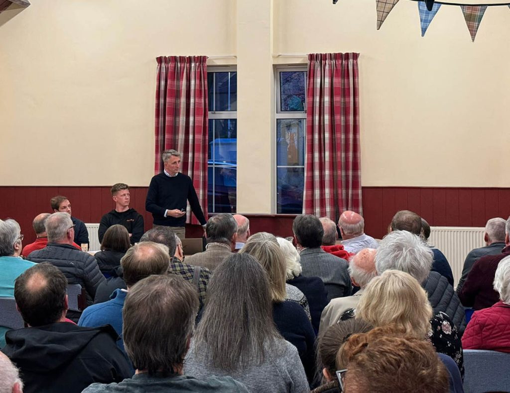 A community meeting for the Eccles Battery Energy Storage System (BESS) project hosted by the Zenobē team, with a speaker addressing a seated audience in a hall decorated with tartan bunting and informational displays on the walls.