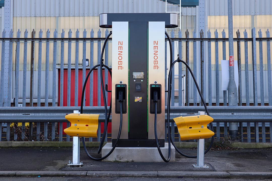 Zerova electric vehicle charging station with Zenobē branding, featuring dual charging ports and protective yellow barriers, installed in an industrial setting with a metal fence in the background.