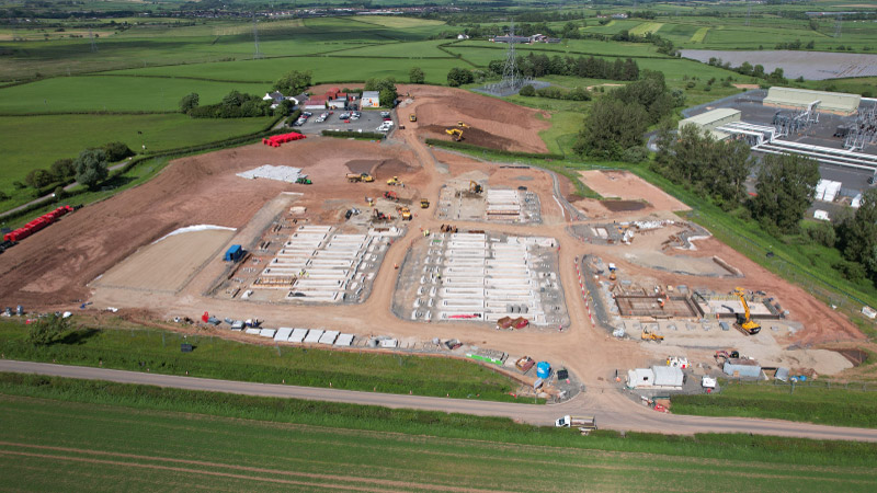 Aerial view of the Kilmarnock South battery storage site under development, showing early earthworks, construction activity, and foundation preparations for large-scale battery installations. Surrounding green fields and a nearby electricity substation are visible in the background. Adjusted for mobile.