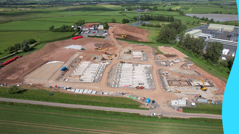 Aerial view of the Kilmarnock South battery storage site under development, showing early earthworks, construction activity, and foundation preparations for large-scale battery installations. Surrounding green fields and a nearby electricity substation are visible in the background.