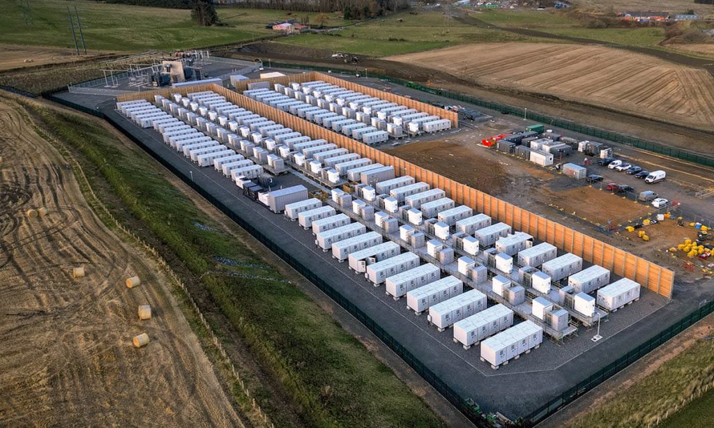 Aerial view of Zenobē’s Blackhillock battery storage site, showcasing rows of white battery containers, wooden wind breaker panels, and surrounding green fields.