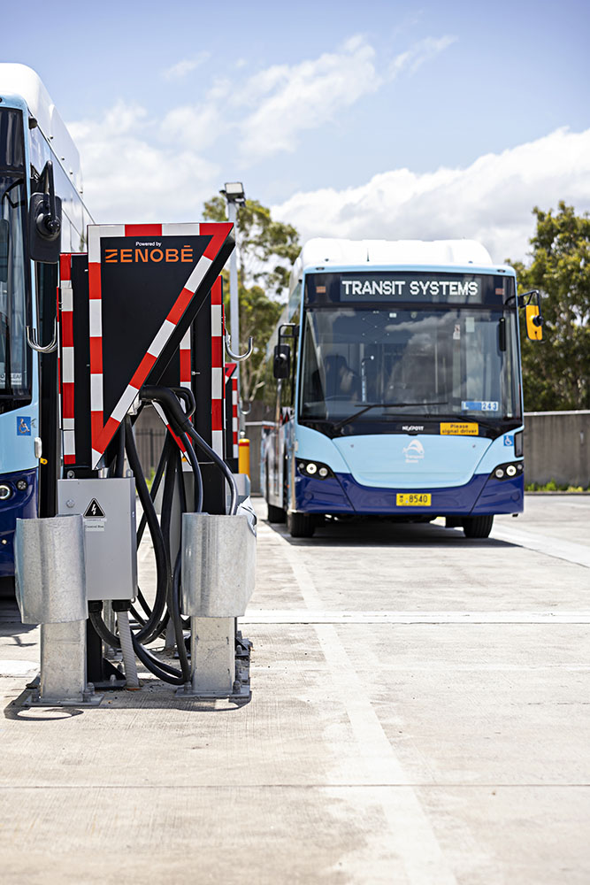 A Zenobē-branded electric bus charger at a Transit Systems depot, with electric buses parked in the background under a bright sky. The charging unit features multiple cables, highlighting the depot’s electric vehicle infrastructure.