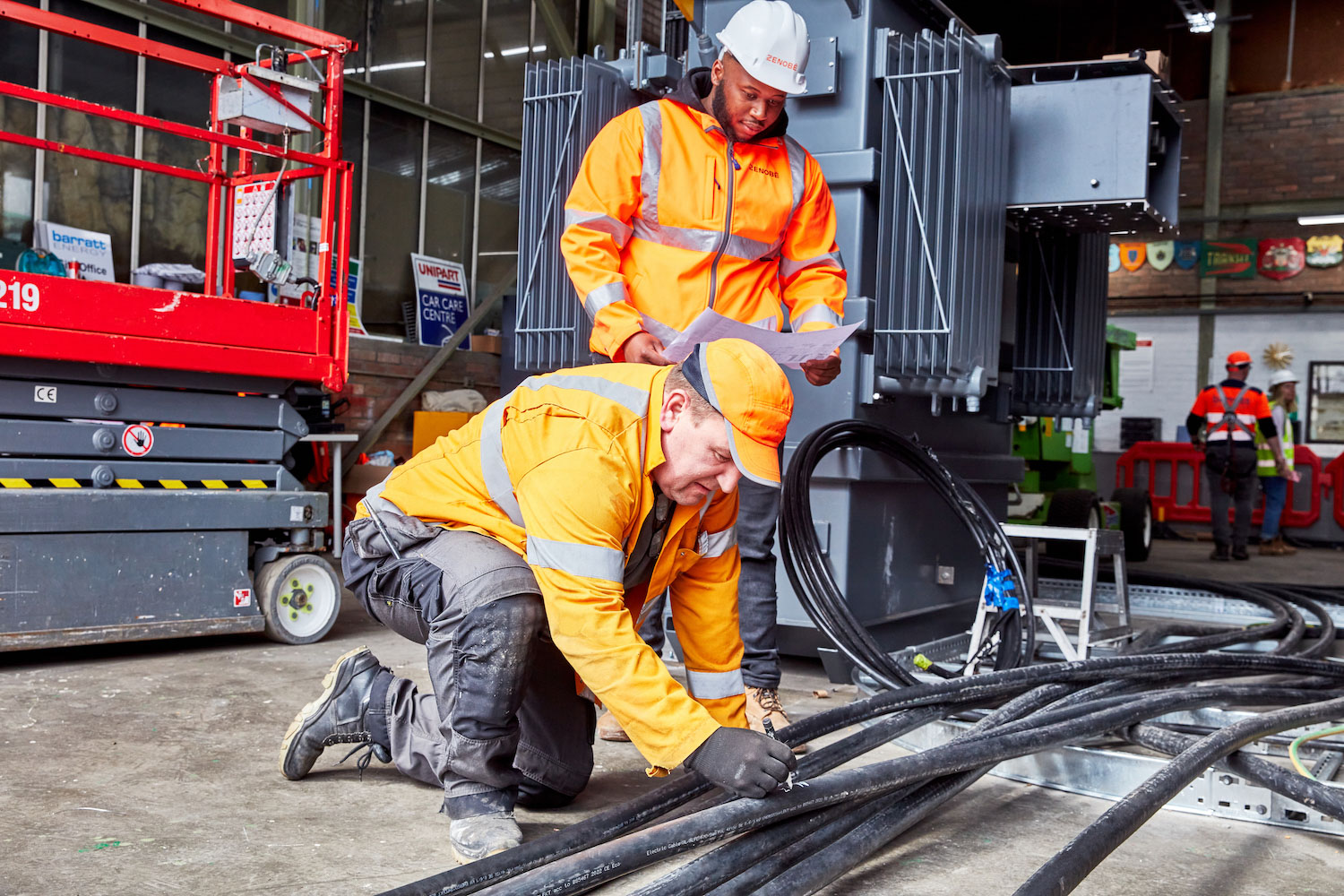 Two men in orange high viz working on a power cable in an electric bus depot
