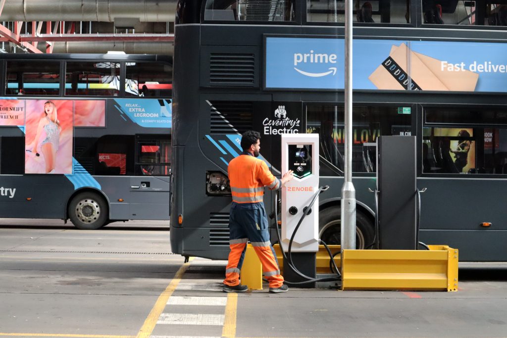Man in orange hi-viz tending to a Zenobe branded EV charger next to a National Express electric bus