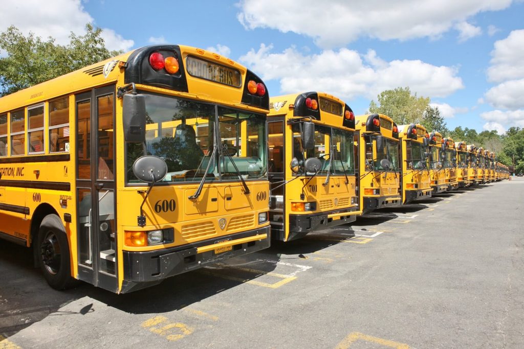 A row of yellow Zenobē-financed electric school buses parked at a depot in New York State, plugged in and charging.